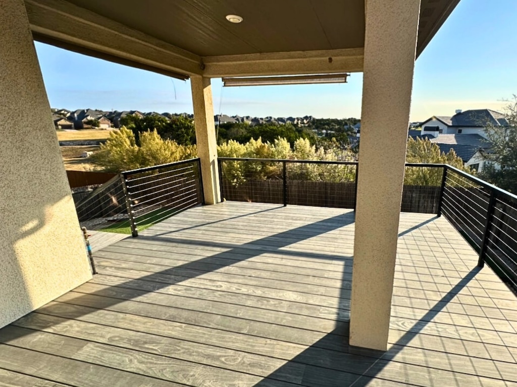 spacious wooden deck with beige pillars and black railing, overlooking a suburban neighborhood and greenery under a clear blue sky at sunset.