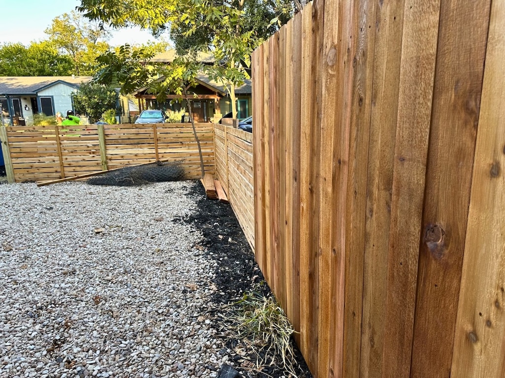 fence-contractors-lakeway wooden fence lines a gravel yard with black mulch. Greenery and houses are visible in the background, creating a peaceful, suburban scene.