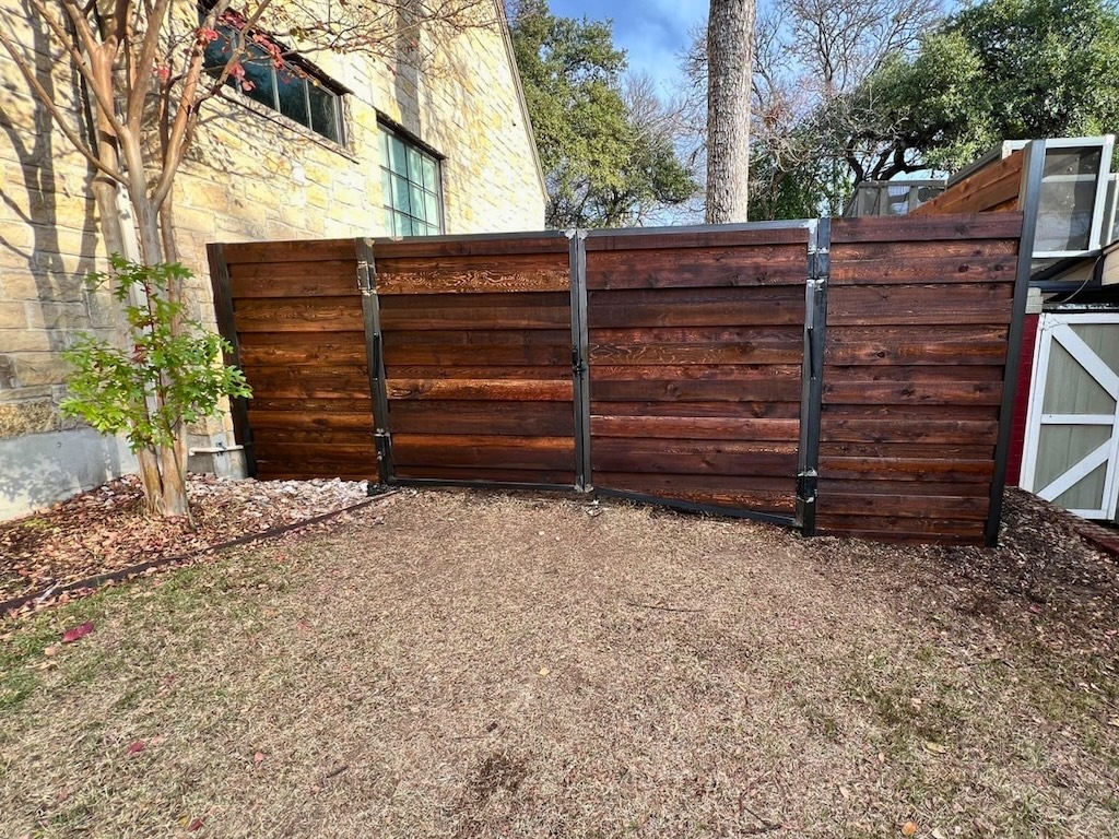 wooden fence with metal hinges against a stone wall near a leafy tree and bare ground. Background features lush trees under a blue sky.