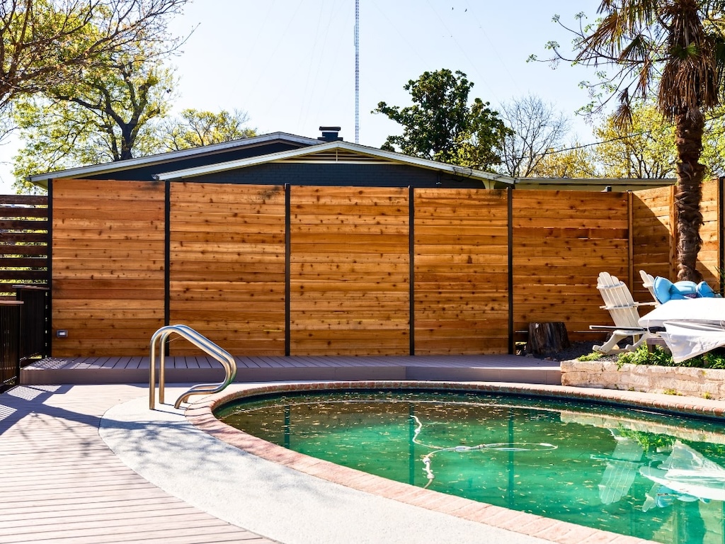 sunny backyard with a curved pool, bordered by a wooden fence. Lounge chairs and a palm tree create a tranquil, inviting atmosphere.