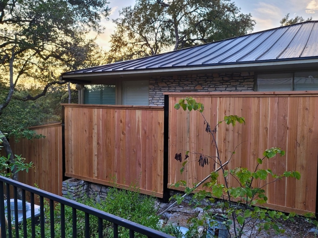 fence-installation-lakeway Wooden privacy fence surrounds a stone house with a metal roof during sunset. Green shrubs and trees create a serene, natural atmosphere.