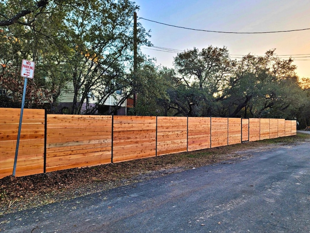 fence-installation-lakeway long, wooden privacy fence runs along a quiet, tree-lined street at sunset. A "No Parking Any Time" sign is visible on the left. The scene feels peaceful.