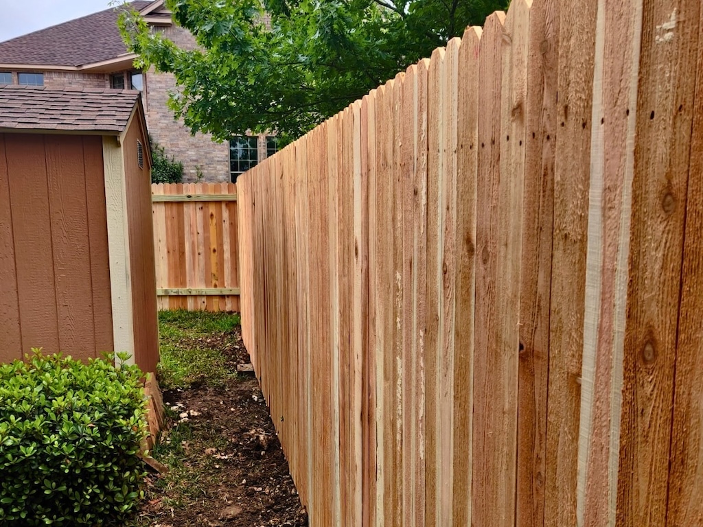 wood-privacy-fence-lakeway new, tall wooden fence runs along a narrow garden path with a small shed and lush green bush on the left. A tree provides a touch of nature.